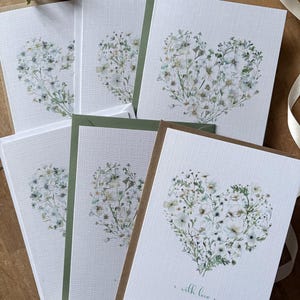 May include: A selection of white greeting cards with a heart-shaped floral design. The cards have a textured surface and are paired with green and brown envelopes. One card is printed with the words "with love".