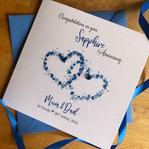 May include: A white card with a blue and white heart design. The card says "Congratulations on your Sapphire Anniversary" and "Mum & Dad 45 Years 29th April 2022".