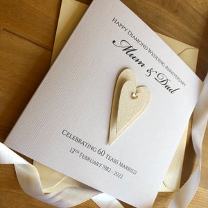 May include: A white card with a textured surface. The card has a black inscription that reads "Happy Diamond Wedding Anniversary Mum & Dad Celebrating 60 Years Married 12th February 1982 - 2022". The card has two layered beige hearts with a sparkly rhinestone in the centre.