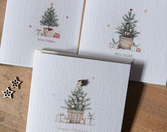 May include: Three square white greeting cards featuring a watercolor illustration of a decorated Christmas tree in a woven basket with gifts. Each card displays the text Merry Christmas or Happy Christmas and includes small wooden star embellishments.