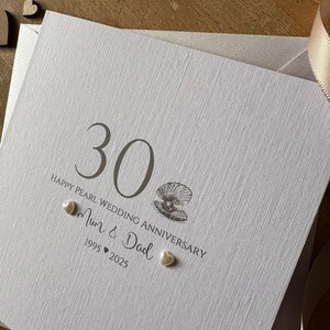 May include: A white textured card with the number 30 and the text "Happy Pearl Wedding Anniversary" printed in gray. The card features a seashell illustration and two heart-shaped pearl embellishments. The card is for a 30th wedding anniversary.