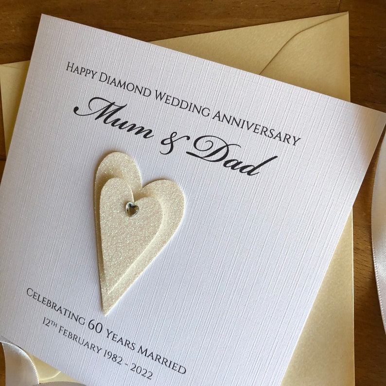 May include: A white card with the text "Happy Diamond Wedding Anniversary Mum & Dad" and "Celebrating 60 Years Married 12th February 1982 - 2022". The card has a sparkly white heart with a small rhinestone in the centre.