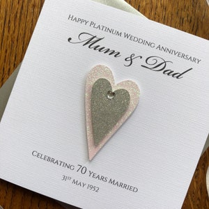 May include: A white card with the text "Happy Platinum Wedding Anniversary Mum & Dad Celebrating 70 Years Married 31st May 1952". The card has two glittery hearts, one pink and one silver, with a rhinestone in the centre.