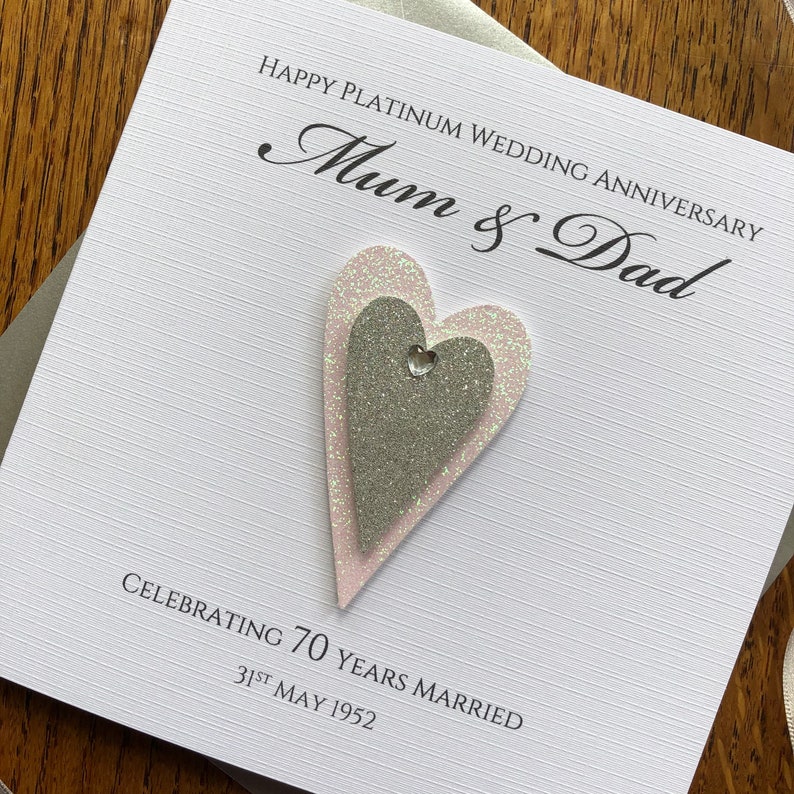 May include: A white card with the text "Happy Platinum Wedding Anniversary Mum & Dad Celebrating 70 Years Married 31st May 1952". The card has two glittery hearts, one pink and one silver, with a rhinestone in the centre.
