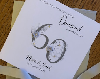 May include: A white card with a gray number 60 decorated with white and blue flowers. The card says "Congratulations on your Diamond Anniversary" and "Mum & Dad 16th June 2022".