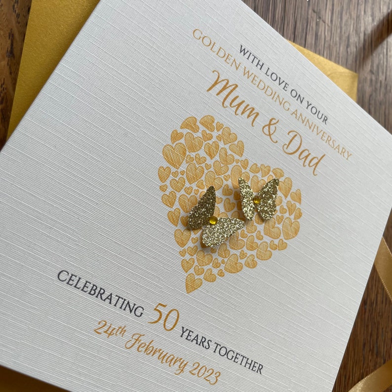 May include: A white card with gold lettering and a gold heart design. The card says "With love on your Golden Wedding Anniversary Mum & Dad Celebrating 50 years together 21st February 2023".