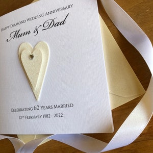 May include: A white card with a glittery heart design and the text "Happy Diamond Wedding Anniversary Mum & Dad Celebrating 60 Years Married 12th February 1982 - 2022"