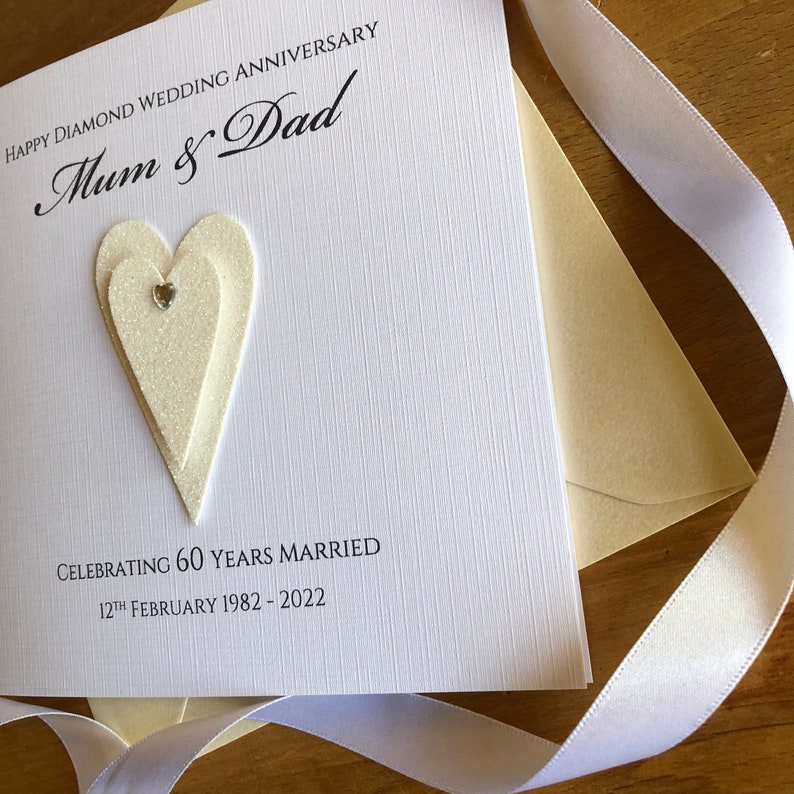 May include: A white card with a glittery heart design and the text "Happy Diamond Wedding Anniversary Mum & Dad Celebrating 60 Years Married 12th February 1982 - 2022"
