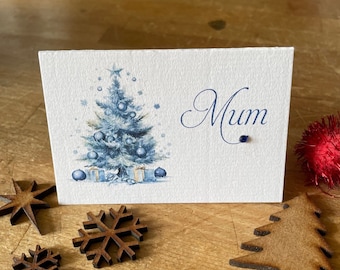 May include: A white Christmas card with a watercolor illustration of a blue Christmas tree, ornaments, and gifts. The word "Mum" is written in elegant blue script. Wooden star and snowflake decorations are scattered around the card.