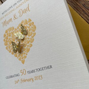 May include: A white card with gold lettering and a gold heart design. The card says "With love on your wedding anniversary, Mum & Dad" and "Celebrating 50 years together, 24th February 2023".