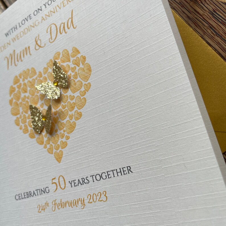 May include: A white card with gold lettering and a gold heart design. The card says "With love on your wedding anniversary, Mum & Dad" and "Celebrating 50 years together, 24th February 2023".