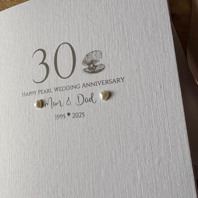 May include: A white card with the text "30" and "Happy Pearl Wedding Anniversary." The card also includes the words "Mum & Dad" and the years "1995 2025." A pearl and shell design is featured.