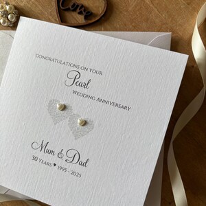 May include: A white Pearl Wedding Anniversary card with the text "Congratulations on your Pearl Wedding Anniversary" and "Mum & Dad". The card features two heart-shaped designs with pearl embellishments and the years 1995-2025.