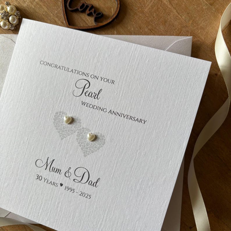 May include: A white Pearl Wedding Anniversary card with the text "Congratulations on your Pearl Wedding Anniversary" and "Mum & Dad". The card features two heart-shaped designs with pearl embellishments and the years 1995-2025.