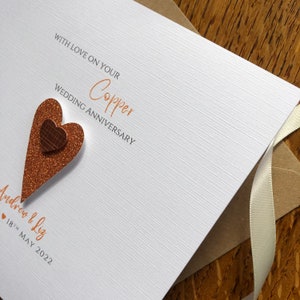 May include: A white card with the text "With love on your Copper Wedding Anniversary" in orange script. The card has a copper glitter heart with a smaller copper heart on top. The text "Andrew & Liz" and "18th May 2022" is written below the heart.