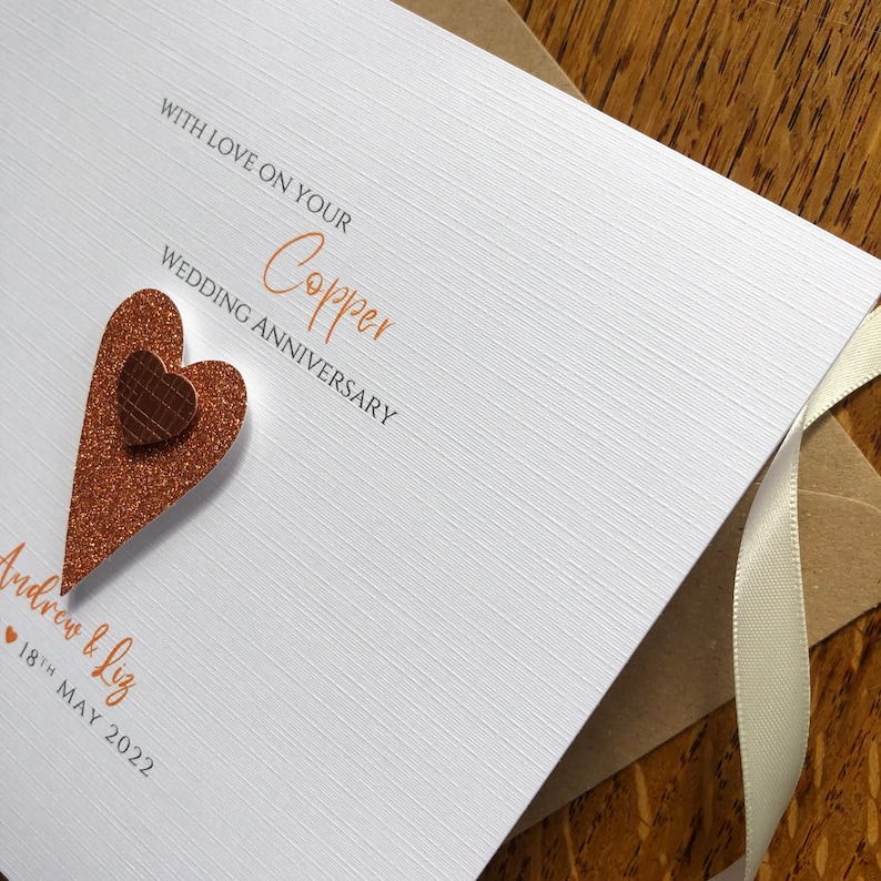 May include: A white card with the text "With love on your Copper Wedding Anniversary" in orange script. The card has a copper glitter heart with a smaller copper heart on top. The text "Andrew & Liz" and "18th May 2022" is written below the heart.