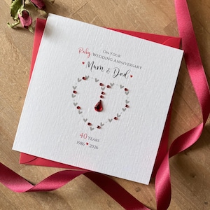 May include: A square white greeting card for a 40th wedding anniversary. The text reads Ruby On Your Wedding Anniversary Mum & Dad 40 Years 1986-2026. A heart shape is formed by small grey hearts and red crystal accents on the front.