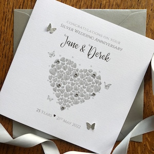 May include: A silver card with a heart made of smaller hearts and the text "Congratulations on your Silver Wedding Anniversary Jane & Derek 25 Years 21st May 2022".