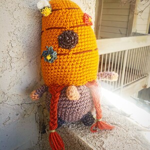 May include: A crocheted gnome toy with a brown and orange hat, a brown beard, and a brown body. The hat has a bee and flower design. The gnome is holding a flower in its hand.