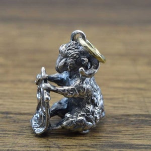 May include: A silver charm shaped like a ram with curly horns. The ram is sitting on its haunches and holding a small, ornate object in its front paws. The charm has a gold loop for attaching it to a necklace or bracelet.