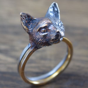以下が含まれることがあります： 猫の頭が細部までデザインされた銀と金のリング。猫の頭は右を向いており、質感のある仕上げになっています。