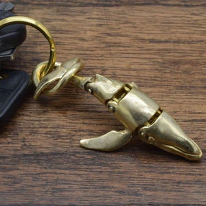 May include: A brass whale-shaped keychain with a gold-colored finish. The keychain features a detailed design of a whale with articulated joints and a sturdy clasp for attaching keys. The whale is resting on a wooden surface.