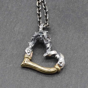 May include: A silver and gold heart-shaped pendant necklace with a detailed, intricate design. The pendant is made of twisted metal and has a polished finish.