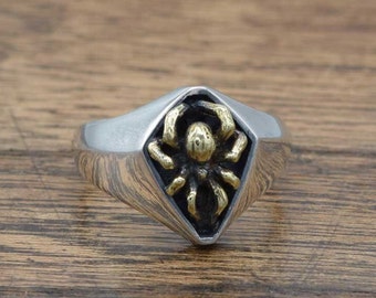 May include: A silver ring with a gold spider design. The ring is shaped like a diamond and has a textured surface.