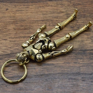 May include: A brass skeleton keychain with a detailed design. The skeleton is lying down with its arms and legs outstretched. The keychain has a circular ring for attaching keys.