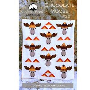 May include: A white quilt featuring a repeating pattern of cartoon moose wearing plaid hats and orange triangles. The quilt has the text "Chocolate Moose #251" and the Cotton Street Commons logo.