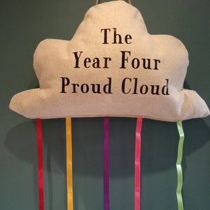 May include: A beige cloud-shaped pillow with the text "The Year Four Proud Cloud" printed on it. Five colorful ribbons hang from the bottom of the pillow.