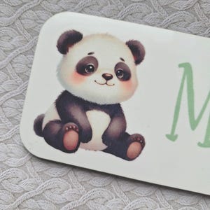 Puede incluir: Letrero rectangular con un oso panda de dibujos animados en blanco y negro, sentado. El panda tiene mejillas rosadas y una expresión sonriente. La letra "M" está escrita en verde en el letrero.