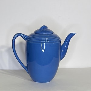 May include: A solid cornflower blue ceramic teapot with a glossy finish. The teapot features a curved handle, a tapered spout, and a circular lid with a small, raised finial handle on top. The body has a smooth, rounded shape with concentric rings.
