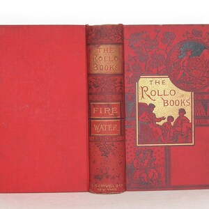 May include: Open antique book titled "The Rollo Books" with red covers and spine. The spine features the title, author, and the words "Fire" and "Water." The right cover has an illustration of people and the title. The book is in good condition.