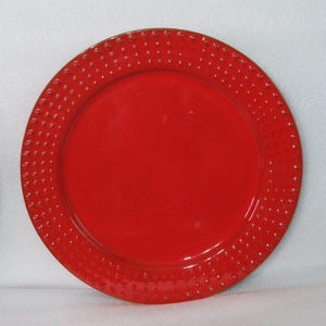 May include: A round, vibrant red dinner plate featuring a smooth, flat center and a wide, textured rim decorated with a repeating pattern of small, raised circular beads that encircle the entire edge of the plate in a uniform, symmetrical design.