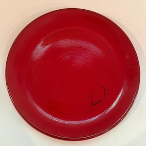May include: A circular plate with a glossy, vibrant red finish. The surface features a thin, black circular line etched toward the center, accompanied by a black handwritten signature located in the lower right quadrant of the plate's interior.