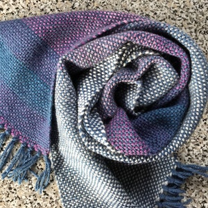 May include: A handwoven scarf in shades of blue, pink, and white with a textured pattern and fringe on the ends.