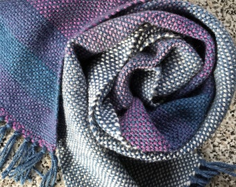 May include: A handwoven scarf in shades of blue, pink, and white with a textured pattern and fringe on the ends.