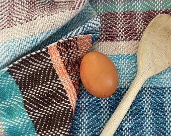 May include: A woven kitchen towel featuring a multi-colored herringbone pattern in shades of blue, brown, orange, and cream. A single brown egg and a light-colored wooden spoon are placed on top of the folded fabric, which shows a textured weave.
