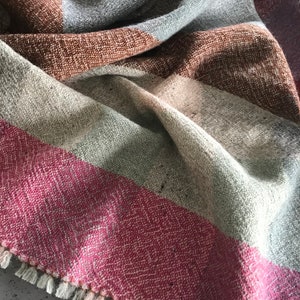 May include: A woven throw blanket with a fringe border. The blanket is made of a soft, textured fabric and features a striped pattern in shades of brown, gray, and pink.
