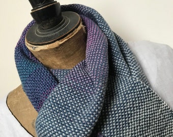 May include: A woven scarf in shades of blue, purple, and white is draped over a mannequin. The scarf has a textured, dotted pattern. The mannequin is brown with a black top. The background is a plain white.