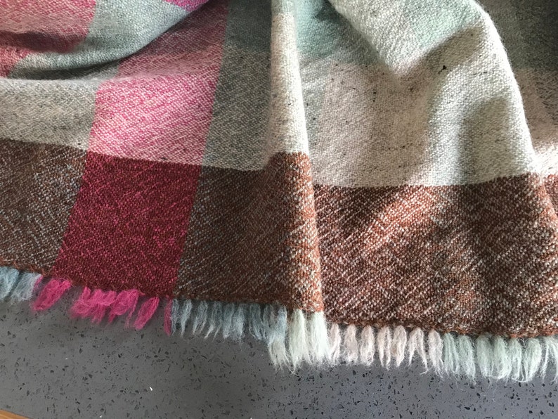 May include: A woven throw blanket with a brown, pink, and gray plaid pattern. The blanket has a fringe border.