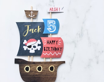 May include: A pirate ship cake topper with a blue and red sail, a skull and crossbones, and the words "Happy Birthday" and "Jack". The ship has a wooden hull with four portholes and a mast with a flag that says "Argh!" and a number "5" on it.