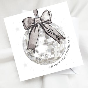 May include: A white square birthday card with a silver disco ball illustration. The card has a silver bow and the name "Brooke" on a ribbon. The text reads "Happy 18th Birthday". The card is on a white background.