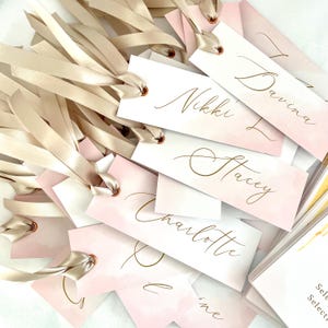 May include: A stack of white gift tags with pink watercolour backgrounds and gold calligraphy names. The tags are tied with beige satin ribbon.