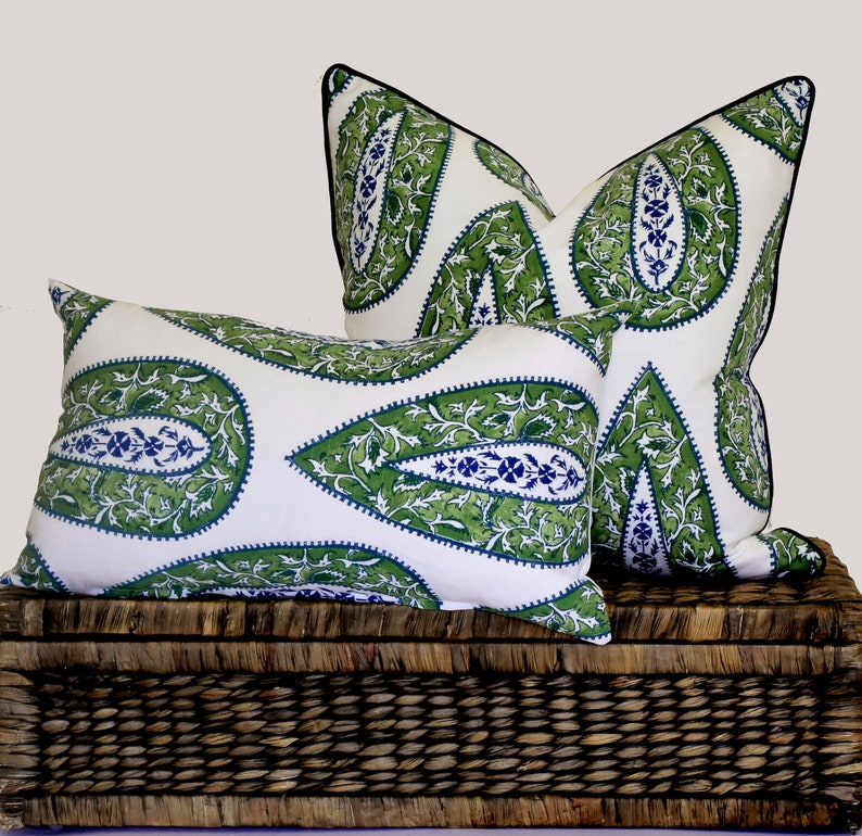 May include: Two decorative throw pillows with a white background and a green and blue paisley pattern. The pillows are sitting on a woven wicker basket.