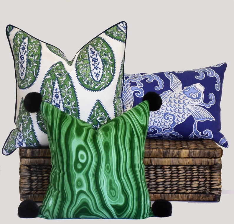 May include: Three decorative throw pillows with pom-poms. The pillows are on a woven wicker basket. The pillows are green, blue, and white with paisley and fish patterns.