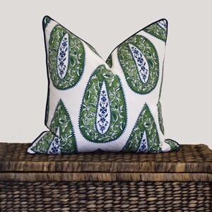 May include: A white pillow with a green and blue paisley pattern. The pillow is sitting on top of a brown wicker storage trunk.