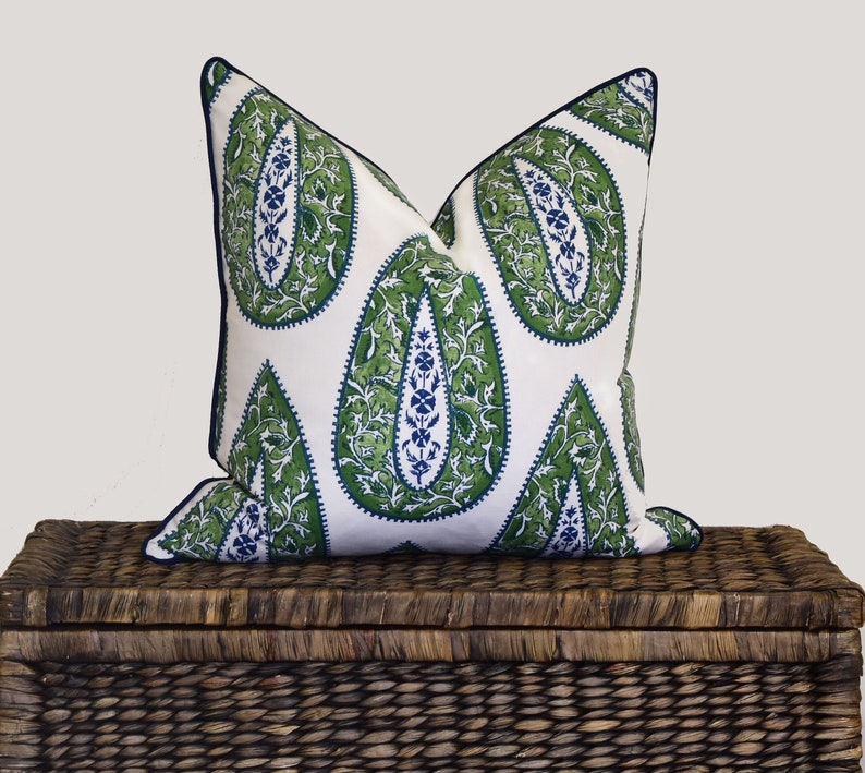 May include: A white pillow with a green and blue paisley pattern. The pillow is sitting on top of a brown wicker storage trunk.