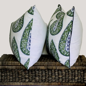 May include: Two white throw pillows with a green paisley pattern and a navy blue trim. The pillows are sitting on a brown wicker basket.
